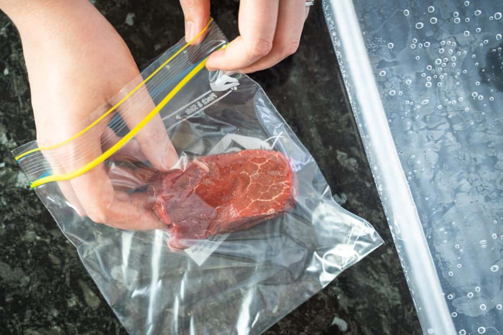 Top view of hands placing a piece of steak inside a zip lock bag, next to a water container.