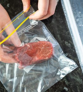 Top view of hands placing a piece of steak inside a zip lock bag, next to a water container.