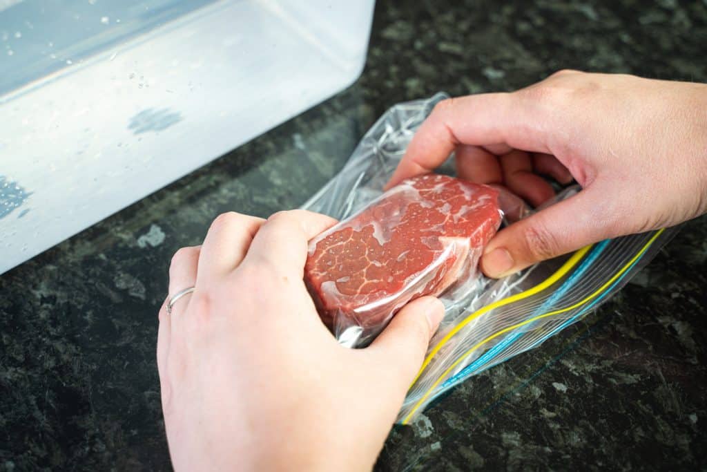 Close-up shot of a piece of steak inside a zip lock bag, with hands squeezing the bag and pushing the air out.