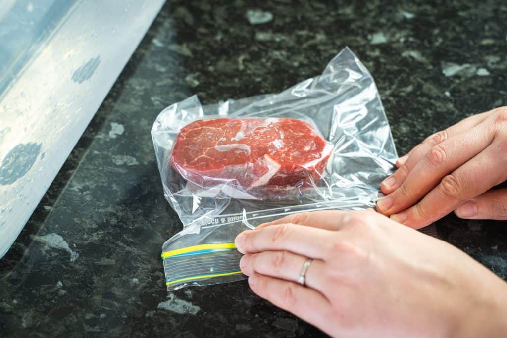 Close-up shot of a piece of steak inside a zip lock bag, with hands closing the bag almost completely, leaving only a small opening at the edge of the bag.