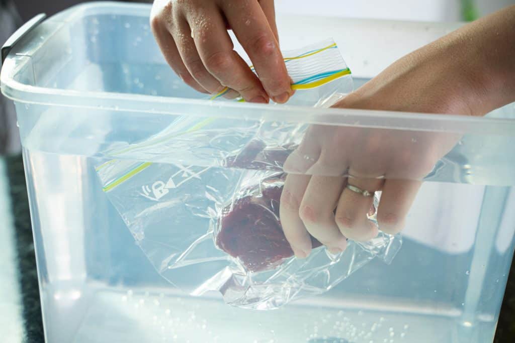 Close-up shot of hands placing a piece of steak inside a zip lock bag inside a container full of water, leaving only an open corner of the bag out of the water.