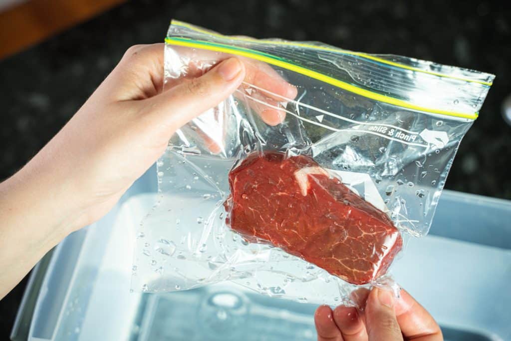 Close-up shot of hands holding a wet, sealed zip lock bag with a piece of steak inside, with a container full of water in the background.