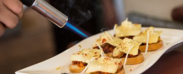 Close up image of a plate with hors d'oeuvre being finished with a kitchen torch that is heating the food.