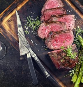Top view of pieces of steak on a wooden chopping board, with herbs and asparagus.
