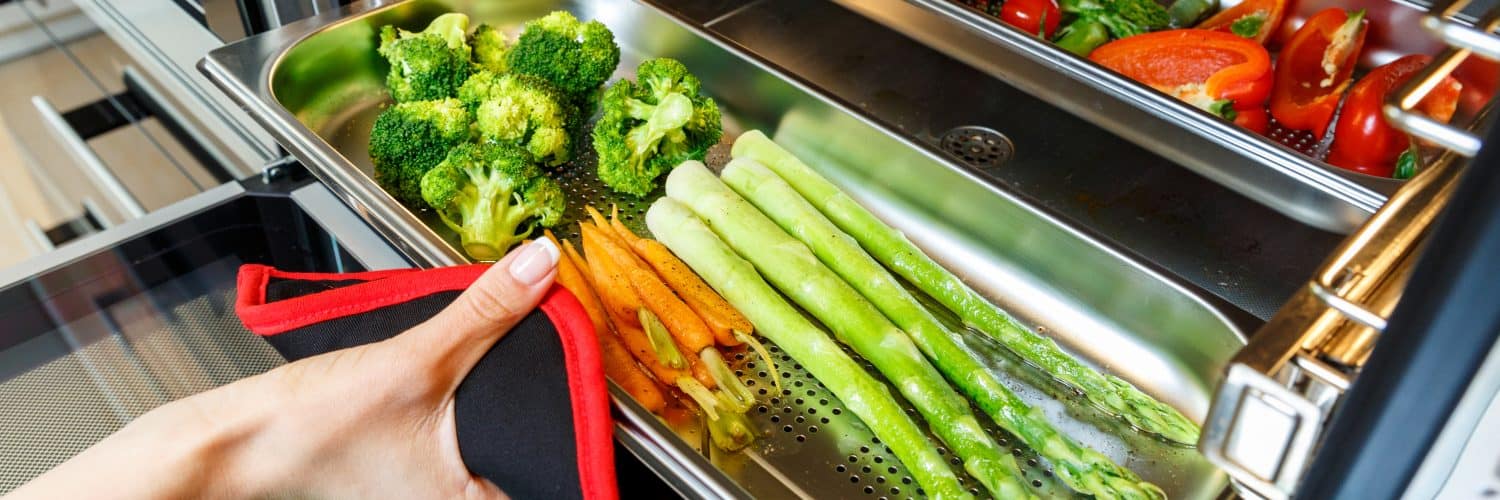 Close-up shot of woman's hand taking a tray full of vegetables out of a steam oven.