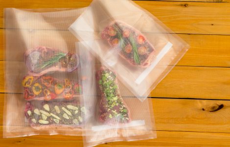 Top view of three vacuum-sealed bags with pieces of steak and herbs on top of a wooden table.