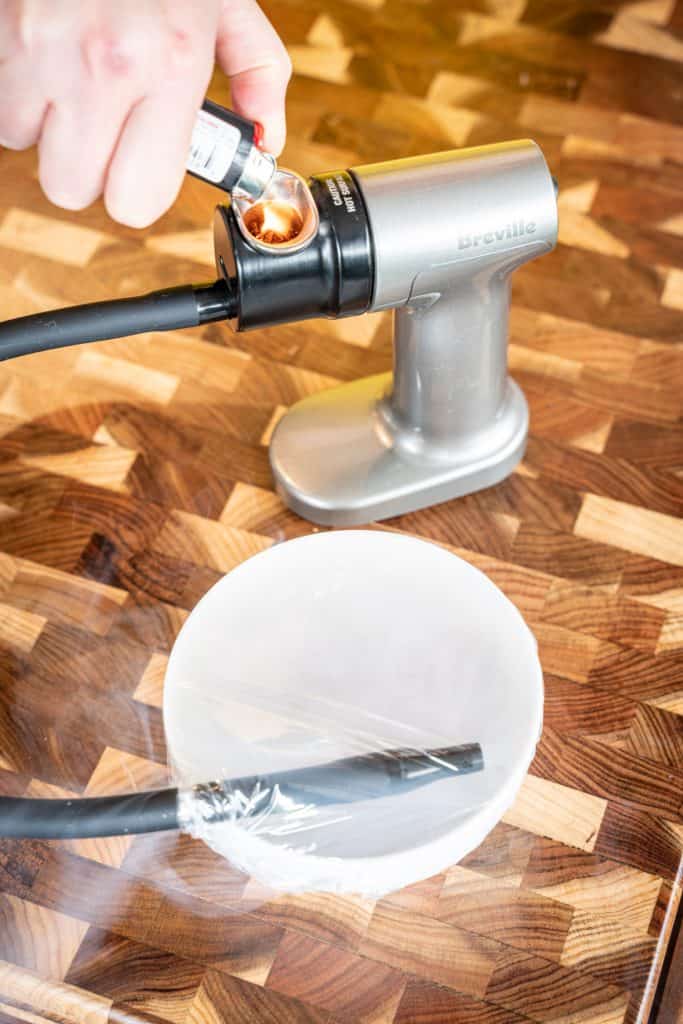 Hand with a lighter lighting up the Breville smoking gun, with hose connecting to bowl full of smoke and topped with plastic wrap.