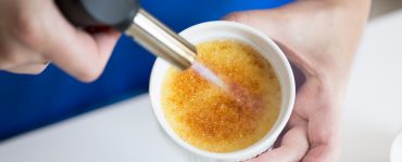 Close-up shot of woman's hand holding a ramekin with creme brulee while using a creme brulee torch to caramelize it.