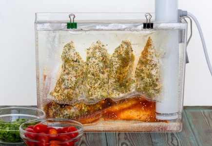 Side view of transparent container full of water, with a vacuum-sealed bag of seasoned chicken submerged and clamped into it, and a sous vide machine.