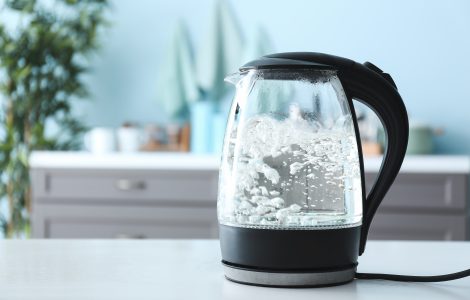 Smart kettle made of glass with boiling water on top of counter and against a blurry kitchen background.