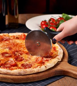 Hand using a pizza wheel to cut a pepperoni pizza placed on a round wooden board