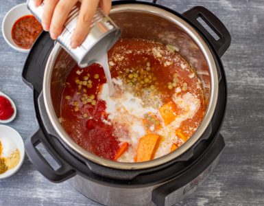 Top view of a slow cooker with sauce and other ingredients in it, with a hand pouring coconut milk from a can inside the pot