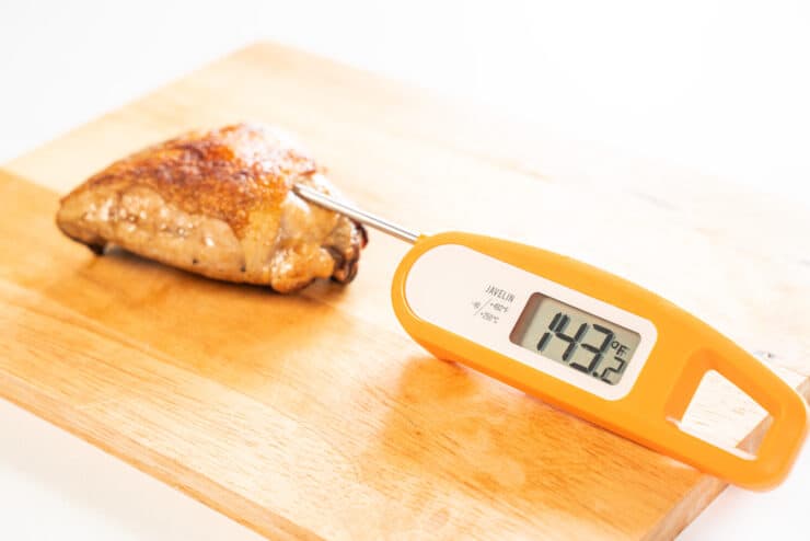 Chicken thigh on cutting board with thermometer inserted into chicken, showing 143.2 degrees Fahrenheit.